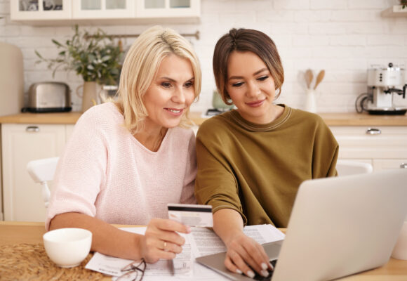 Happy mature blonde woman holding plastic card sitting in front of open laptop with her young daughter, shopping online, using banking application to make payment. E-commerce, internet and technology Mulheres respondem por 65% dos pedidos de crédito no varejo, aponta levantamento